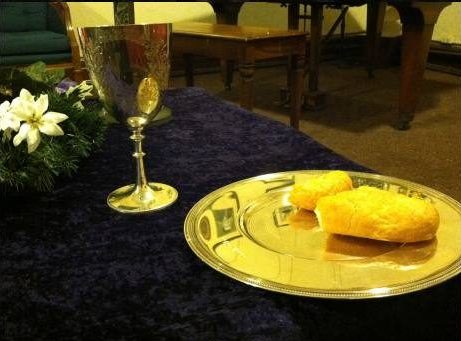 Silver Communion chalice and plate with bread. There is an articifial flower to the left of the chalice, and it all sits on a dark velvet cloth.