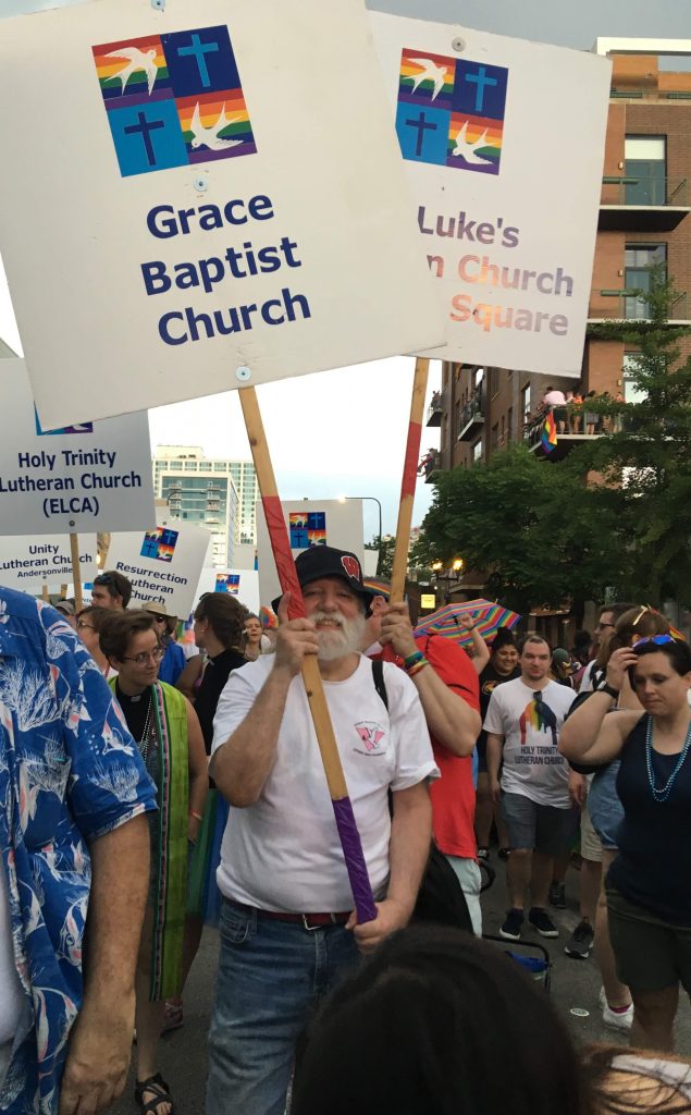 A white man with a white beard, a friendly smile, white Grace Baptist Church T-shirt, jeans, and a black hat holds a sign that says "Grace Baptist Church" in a crowd of people at a Pride Parade.